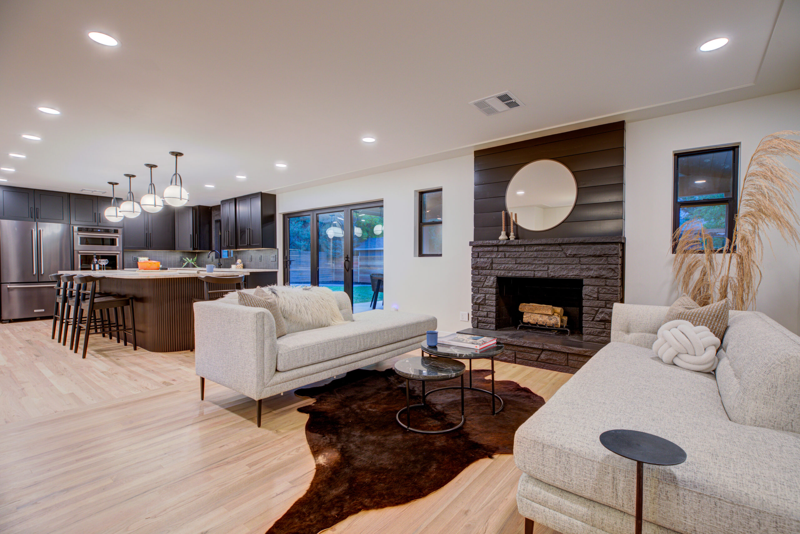 A modern, open-concept living room in a Denver home with a cozy seating area, fireplace, and integrated kitchen, featuring clean lines and a warm color palette. It includes natural light streaming in through